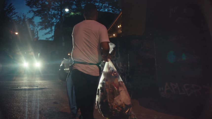 A restaurant worker throws a large bag of trash into a graffiti-covered dumpster in a dark urban alley at night