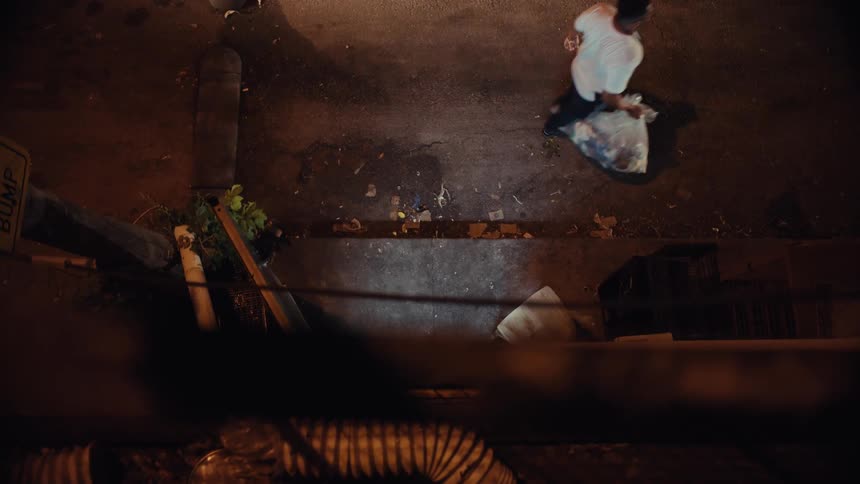 A high-angle view shows a person carrying a flashlight and a trash bag through a dark, dirty urban alley at night