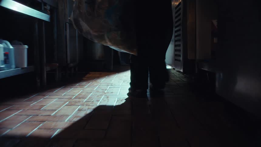 A person drags a large, dripping trash bag across the wet floor of a dark commercial kitchen in slow motion