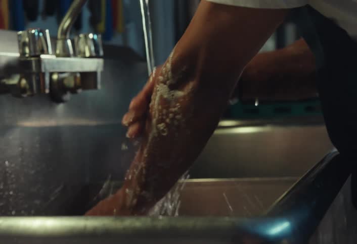 A male chef meticulously washes his soapy hands and arms under running water in a commercial kitchen sink