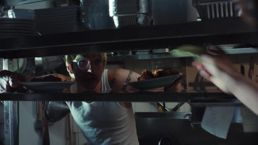 A waiter takes finished dishes from a pass-through shelf as a chef works in a busy restaurant kitchen