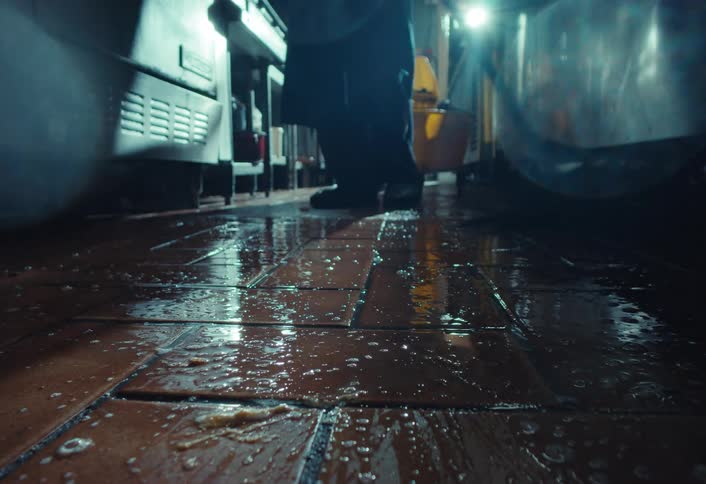 Slow motion, low-angle shot of a restaurant worker mopping the dirty tiled floor of a commercial kitchen