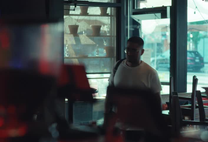 A man in a white t-shirt and glasses walks into a dimly lit bar from the bright street outside and greets a person at the counter