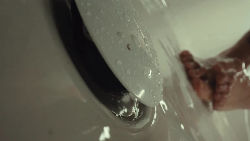 Slow motion close up of water draining into the overflow of a bathtub while a person washes their feet in the background