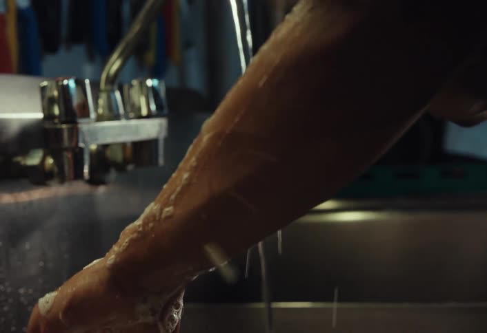 A worker in an apron meticulously scrubs their hands and forearms with a bar of soap in a stainless steel sink