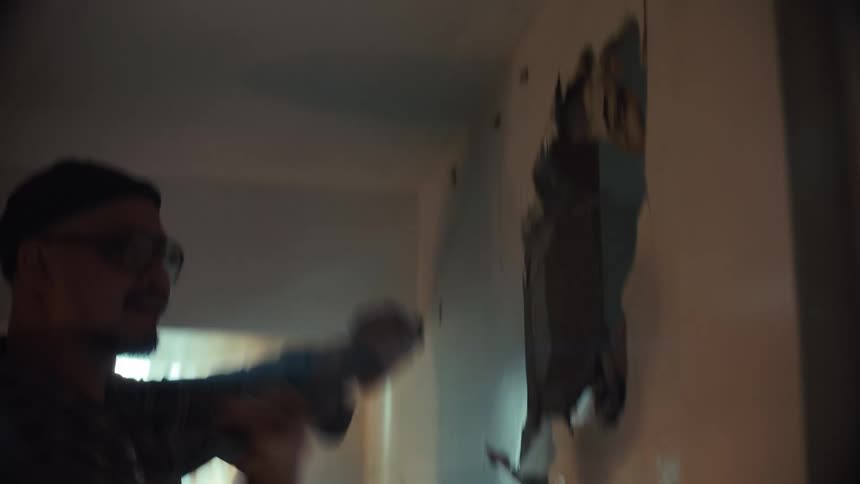 Intense close-up of a man wearing glasses and a beanie furiously demolishing a drywall with a hammer in a dusty room