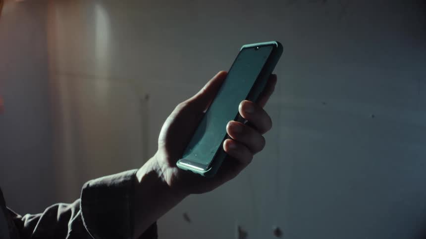 A close-up shot shows a hand holding a smartphone in a dimly lit room with dramatic lighting