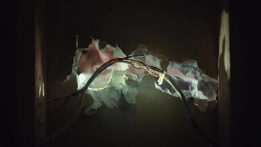 View from inside a dark wall as a person shines a flashlight and peers through a jagged hole, their eye scanning the hidden space