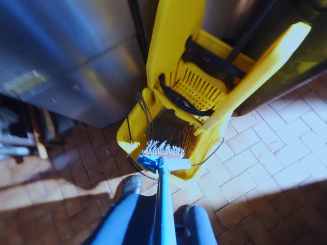 A first-person point of view of mopping the tile floor of a professional kitchen and wringing out the mop in a yellow bucket