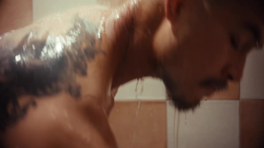 A tattooed man applies body wash to his hands in a steamy shower in slow motion
