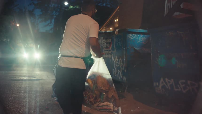 A restaurant worker in an apron takes out a large bag of garbage and throws it into a dumpster in a gritty urban alley at night, filmed in slow motion