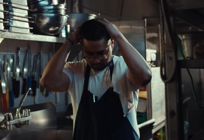 A male chef styles his hair with pomade in a busy restaurant kitchen before feeling stressed and exhausted