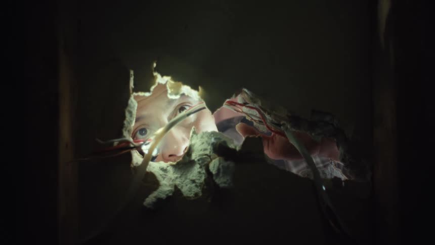 A curious man with a headlamp shines a flashlight into a dark, dusty hole in the wall to inspect the wiring inside