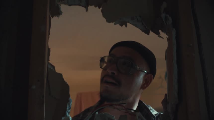 A handyman wearing glasses and a beanie peers through a hole in a drywall during a home renovation project