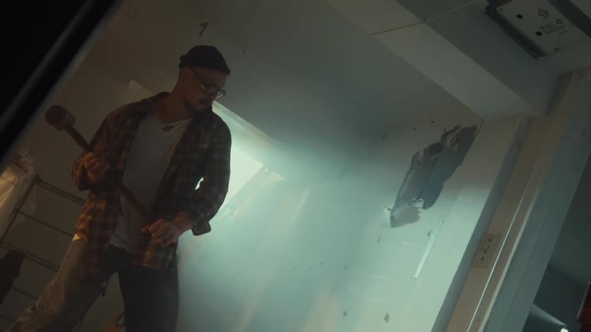 A determined man in a dusty room slowly raises a large sledgehammer to demolish a wall