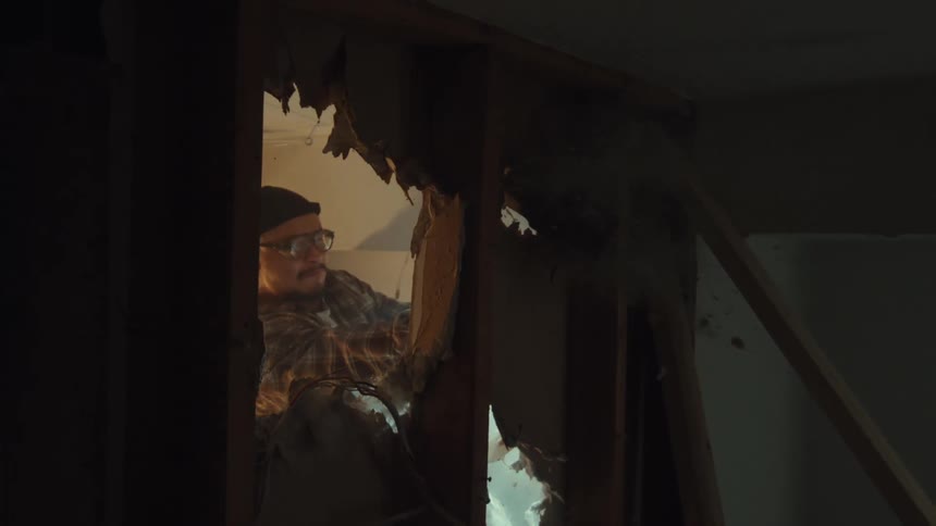 Slow motion shot through a hole in the wall captures a determined man demolishing drywall with a sledgehammer during home renovation