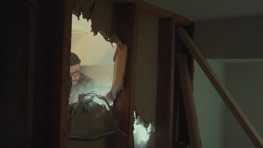 A construction worker in a beanie and glasses is framed by a demolished wall while using a power tool, with dust particles floating in the air in slow motion