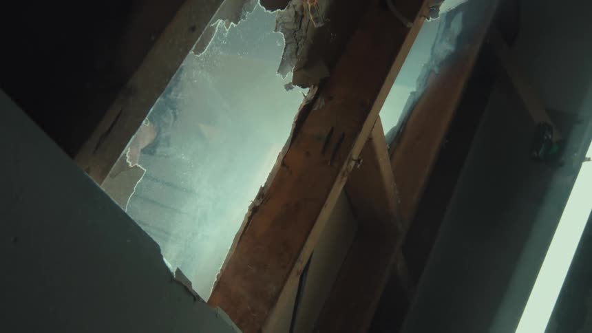 From a low-angle, slow-motion perspective, a person uses a sledgehammer to break through a wooden floor, sending dust and debris raining down