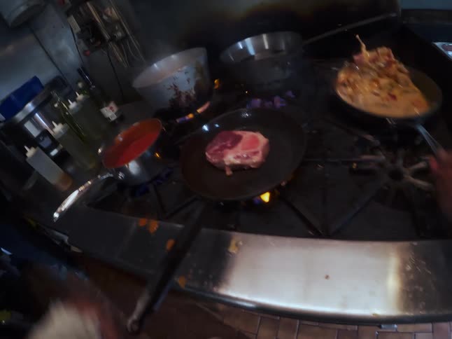 First-person perspective of a professional chef multitasking by searing a steak and preparing sauces on a busy restaurant stove