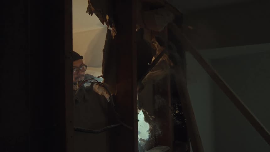 A construction worker demolishes a wall in a dark room in dramatic slow motion, backlit by a bright light