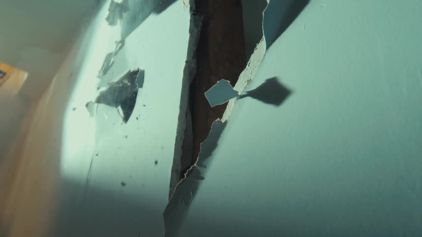 Dust and debris fall in slow motion from a gaping hole in a damaged interior drywall