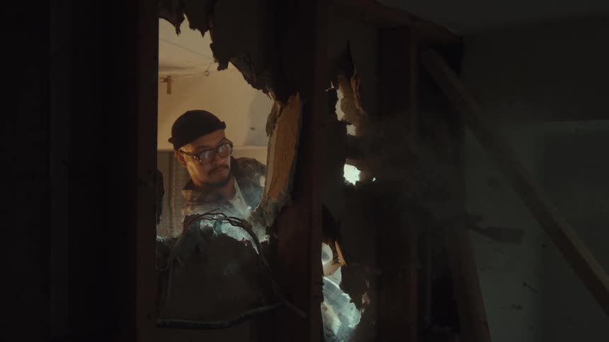 A man wearing glasses and a beanie peers through a hole in a wall as debris flies in slow motion during a demolition
