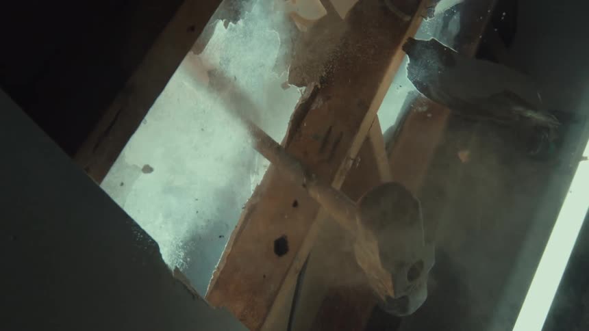 A construction worker smashes through a wooden ceiling with a sledgehammer in dramatic slow motion, viewed from below