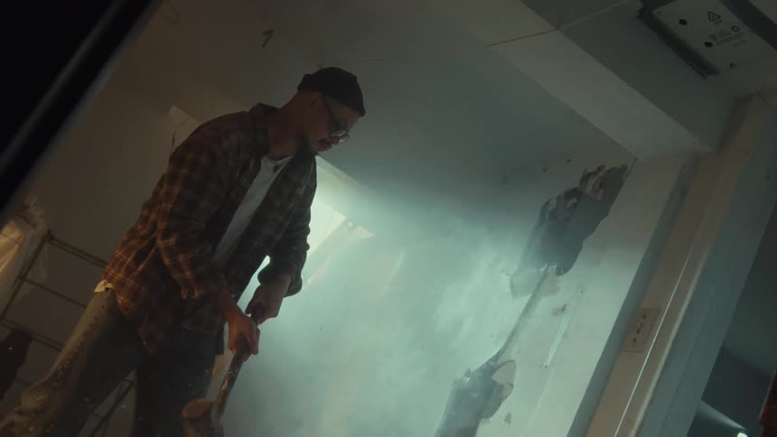 A man wearing a plaid shirt smashes a drywall with a sledgehammer in dramatic super slow motion