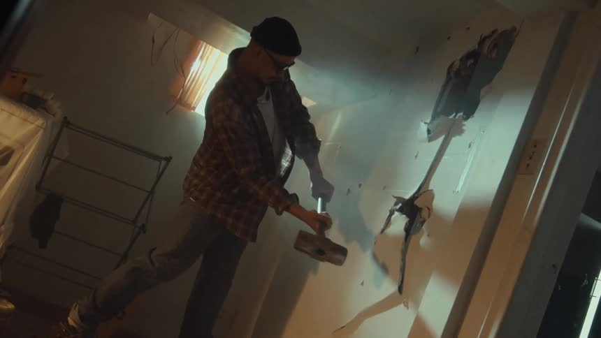 A man in a beanie and plaid shirt powerfully swings a sledgehammer in dramatic slow motion, demolishing a drywall in a dusty room