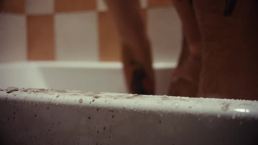 A close-up of a person with tattoos washing their legs in a dimly lit bathtub