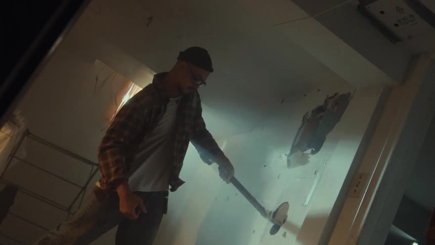 Cinematic slow motion footage shows a man breaking down a drywall with a sledgehammer during a home renovation
