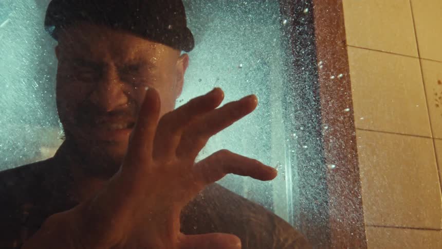 Slow motion close-up of a distraught man in a beanie under a shower, his hand slowly reaching for the glass door