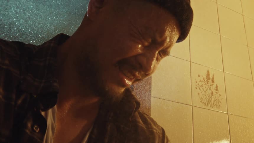 A distraught and emotional man in a beanie cries in a tiled shower with his clothes on, overwhelmed with sadness and despair in slow motion