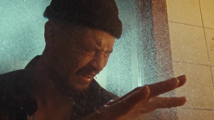A distraught man with a beanie endures a powerful shower spray in a dramatic, slow-motion close-up