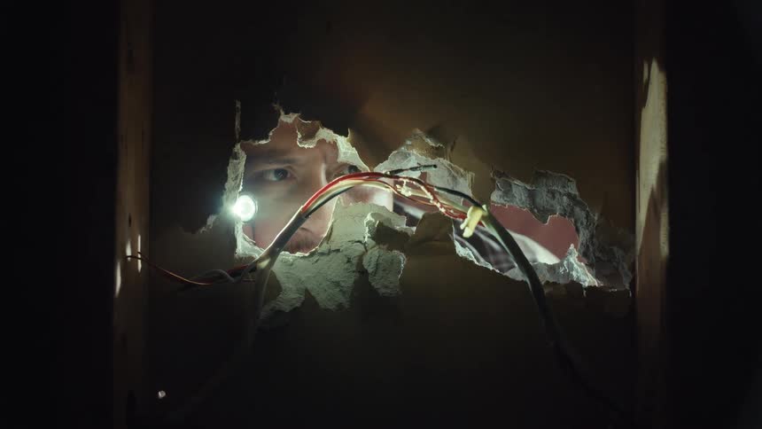 An electrician with a headlamp shines a light through a hole in the wall, looking for the source of an electrical problem
