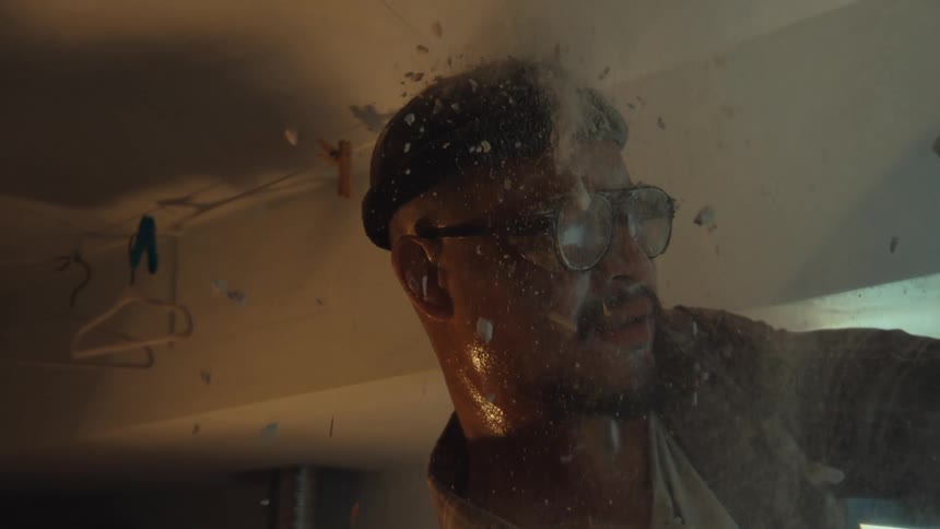A sweaty man in glasses and a beanie watches in shock as debris and dust fall on him in dramatic slow motion