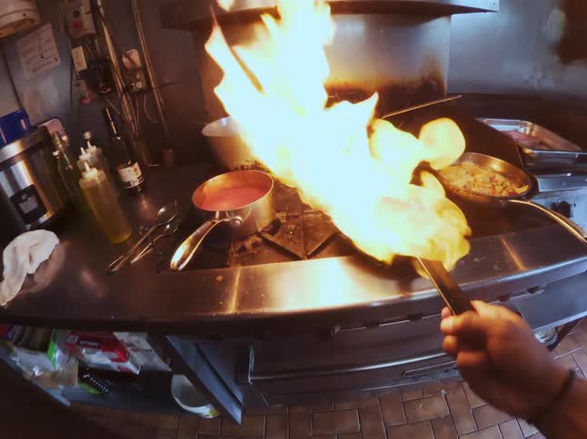 A first-person view of a chef expertly flambéing a steak in a sizzling pan in a restaurant kitchen
