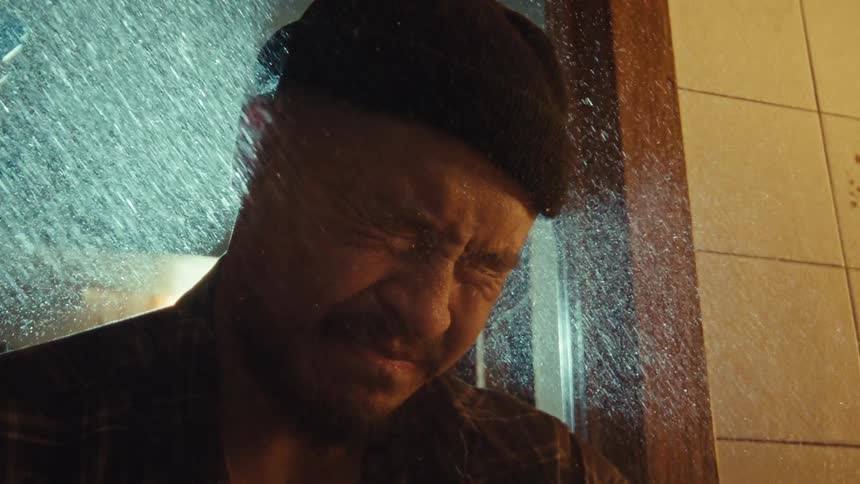 A distressed man in a beanie gets sprayed with water in slow motion, expressing deep emotional turmoil
