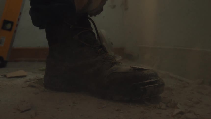 Debris and dust from a demolished wall falls in slow motion around a construction worker's boot