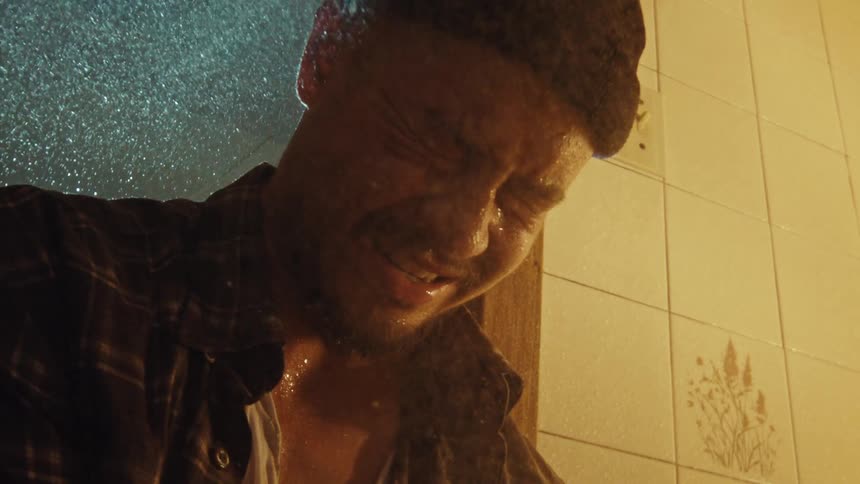 An emotional man in a beanie and plaid shirt cries with his eyes shut as water pours over him in a shower in slow motion