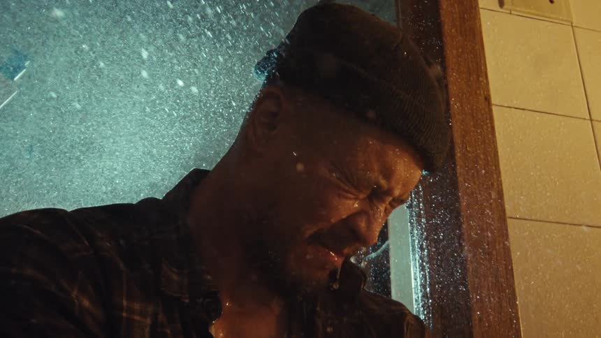 Slow motion footage of a man with a beanie getting drenched in the rain, his face showing deep anguish
