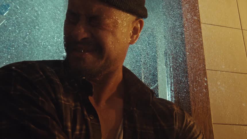 A distraught man in a beanie and plaid shirt is drenched by a heavy spray of water in a dramatic slow-motion close-up