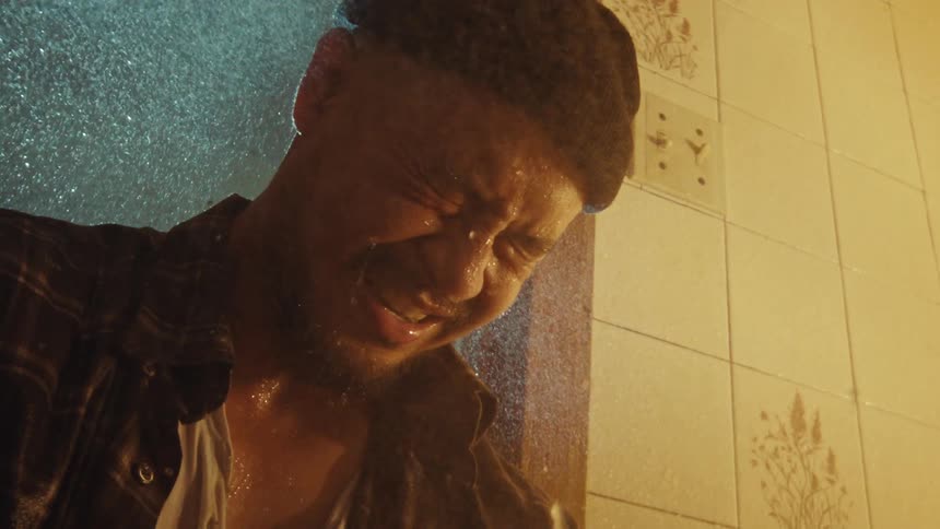 Slow motion close-up of a tormented man in a beanie grimacing in pain as water pours down on him