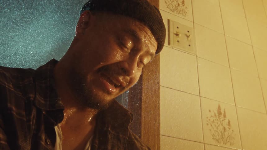 An emotional man in a beanie leans against a tiled wall, drenched by a shower in slow motion