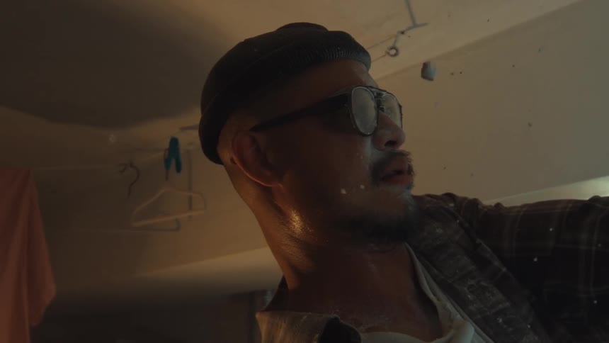 A sweaty man in a beanie watches in slow motion as dust and debris from a crumbling ceiling fall onto his face