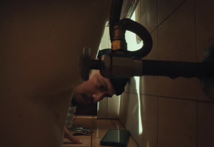 A focused plumber lies on a tiled bathroom floor examining pipes, using his smartphone as a light source