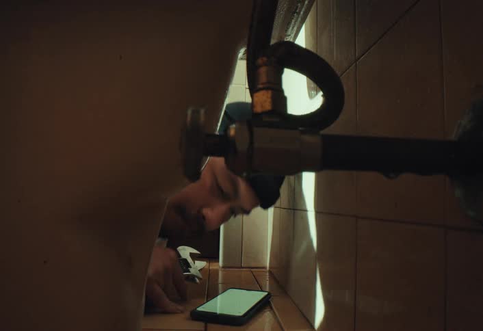 A plumber in a black beanie uses his smartphone for guidance while repairing a leaky pipe in a tight bathroom space
