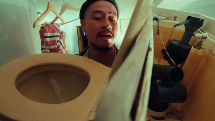 A man wearing a beanie looks down into a dirty toilet with a horrified expression, shot from a POV inside the bowl