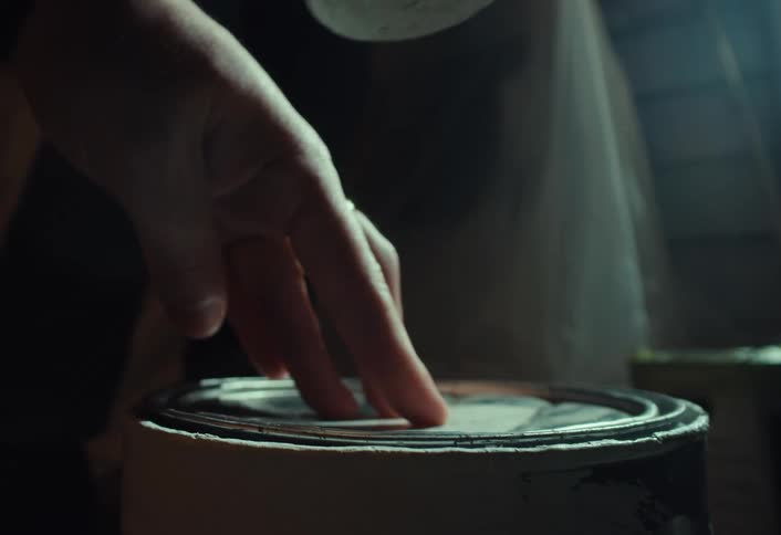 Close up of a person using a rubber mallet to hammer shut the lid of a paint can in a dimly lit workshop