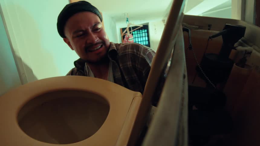 A stressed man grimaces in frustration as he struggles to fix a broken toilet, seen from a low angle